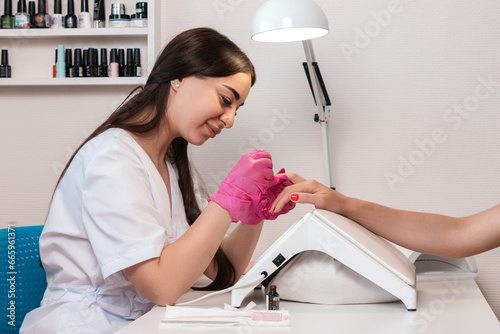 Pretty caucasian manicurist in pink medical gloves paints the client's nails with bright pink varnish. Indoor. The concept of a professional care at manicure salon