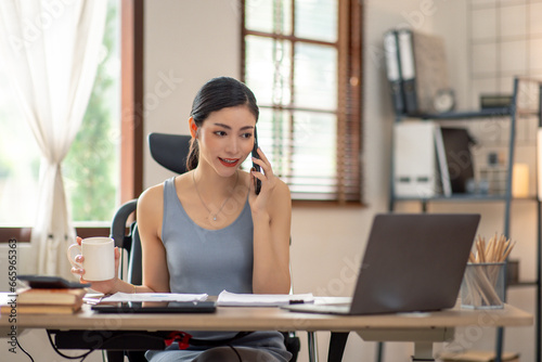 Asian Woman entrepreneur busy with her work in the office. Young freelancer Asian woman talking over smartphone or cellphone while working on computer at her desk.