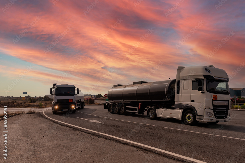 Two tankers with dangerous goods, one parked and one driving under a dramatic sky at dawn.