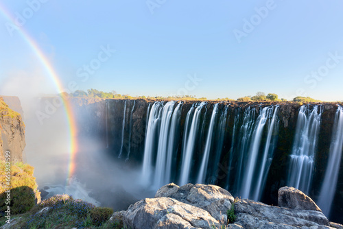 Panorama photo of Victoria Falls waterfall on Zambezi river in very high flow in late evening light with rising spray and intensive rainbow over falls.