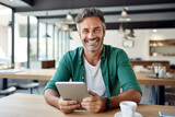 Cheerful Caucasian middle-aged man in casual clothes holding a tablet sits at table in cafe. Happy smiling mature businessman, successful entrepreneur or employee works online. Remote work concept.