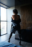 Beautiful tall brunette woman with short hair in black leather coat show her muscular shoulders and back. Tanned skin and black leather   knee-high boots on heels. Bedroom and window