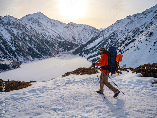 A man traveler makes trekking high in the mountains in winter. A man in a red down jacket with a backpack walks against the background of a frozen mountain lake