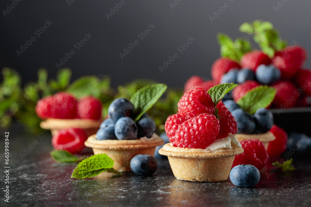 Small tartlets with fresh raspberries and blueberries on a black background. - obrazy, fototapety, plakaty 