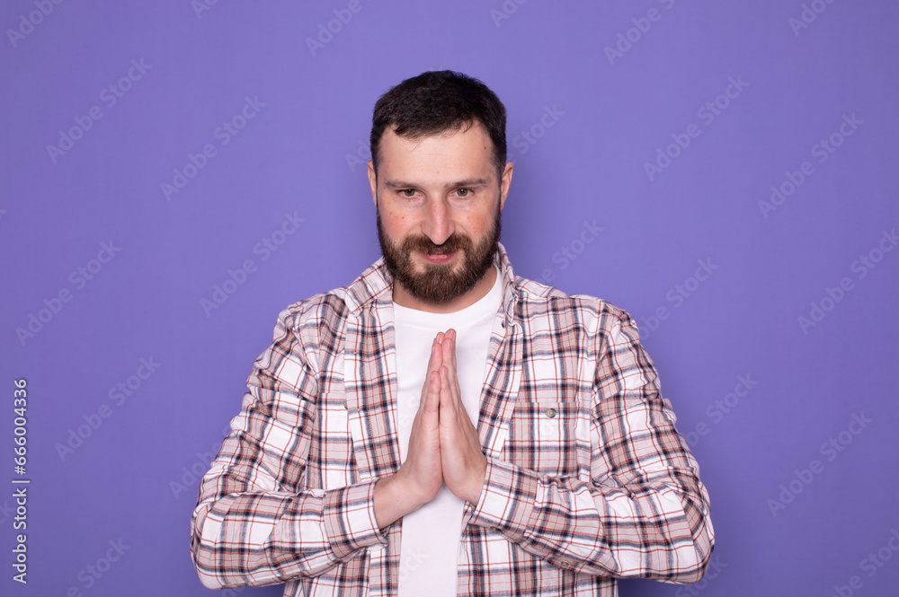 Young bearded caucasian man isolated on purple background holding hands in pray near mouth, feels confident. 