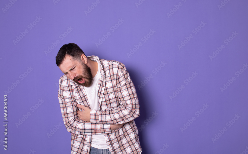 Young bearded man having a stomachache. Young man keeping hands at his stomach because painful disease feeling unwell on purple background. Ache concept. Young bearded man having a stomachache. Young man keeping hands at his stomach because painful disease feeling unwell on purple background. Ache concept.