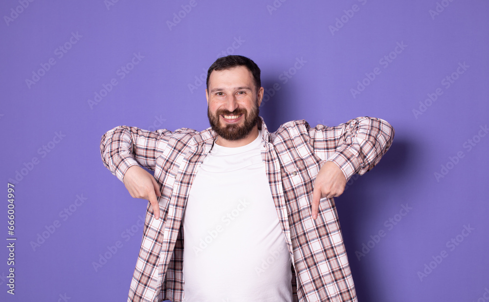 Young bearded man isolated on purple background points down with fingers, positive feeling.