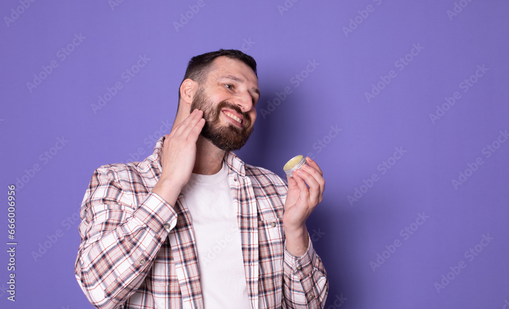 Handsome bearded man holding beard balm on purple background. Handsome bearded man holding beard balm on purple background.
