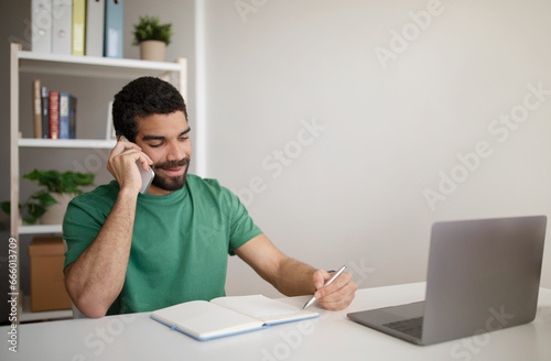 Cheerful millennial arab man manager at table with laptop working remotely, call by phone