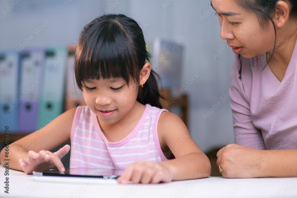 Happy little Asian daughter talking with mother using digital tablet to do homework or study online. Happy young girl and adult sitting at table learning on iPad together. Back to school. Happy little Asian daughter talking with mother using digital tablet to do homework or study online. Happy young girl and adult sitting at table learning on iPad together. Back to school.