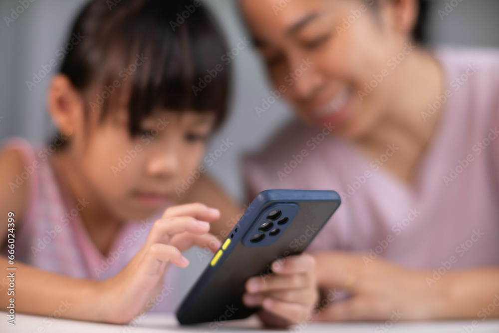 Fototapeta premium Happy Asian family mother and cute daughter using phone together at home. Mother and little girl laughing happily using smartphone watching funny social media video app sitting at table together.