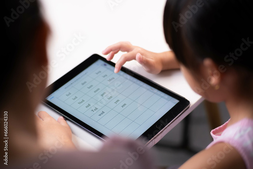 Happy little Asian daughter talking with mother using digital tablet to do homework or study online. Happy young girl and adult sitting at table learning on iPad together. Back to school.
