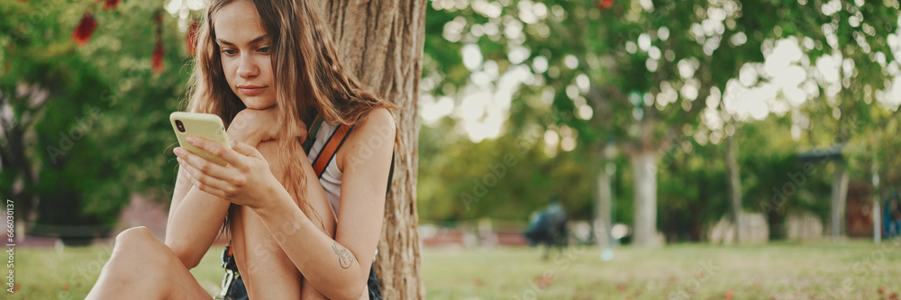 Beautiful girl with long wavy hair wearing in white top uses mobile phone while sitting on the green grass under tree in the park. Girl flip through the pictures on your phone with fingers Beautiful girl with long wavy hair wearing in white top uses mobile phone while sitting on the green grass under tree in the park. Girl flip through the pictures on your phone with fingers