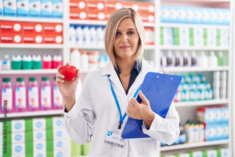 Young woman working at pharmacy drugstore holding heart relaxed with serious expression on face. simple and natural looking at the camera.