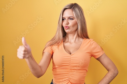 Young woman standing over yellow background looking proud, smiling doing thumbs up gesture to the side