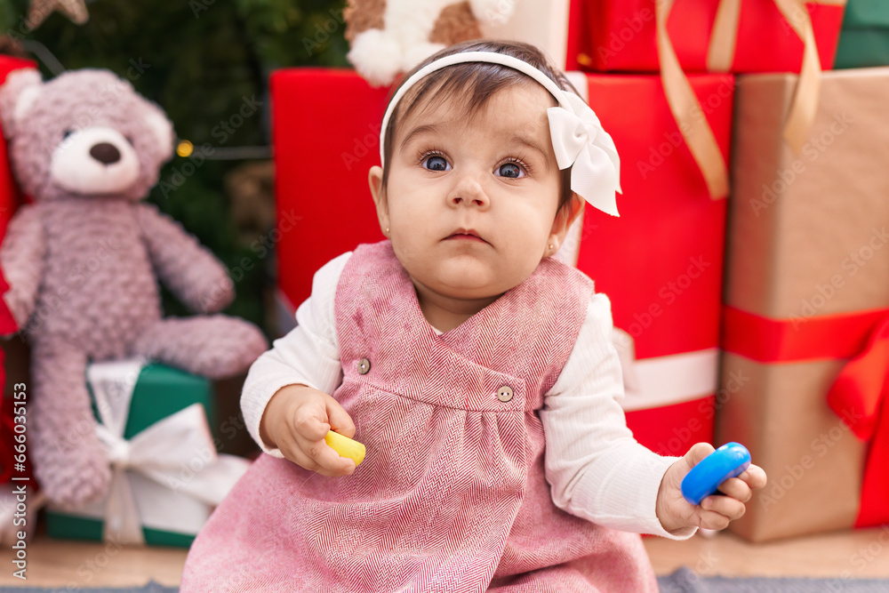 Adorable hispanic baby holding hoops toy sitting on floor by christmas tree at home Adorable hispanic baby holding hoops toy sitting on floor by christmas tree at home