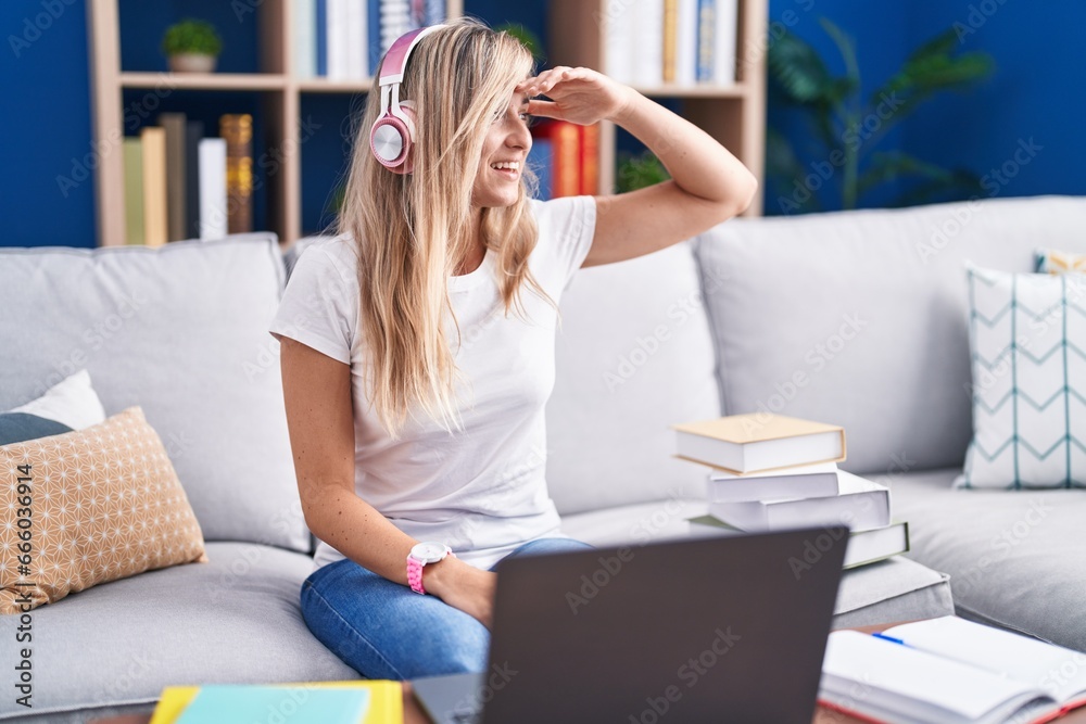 Young blonde woman studying using computer laptop at home very happy and smiling looking far away with hand over head. searching concept.