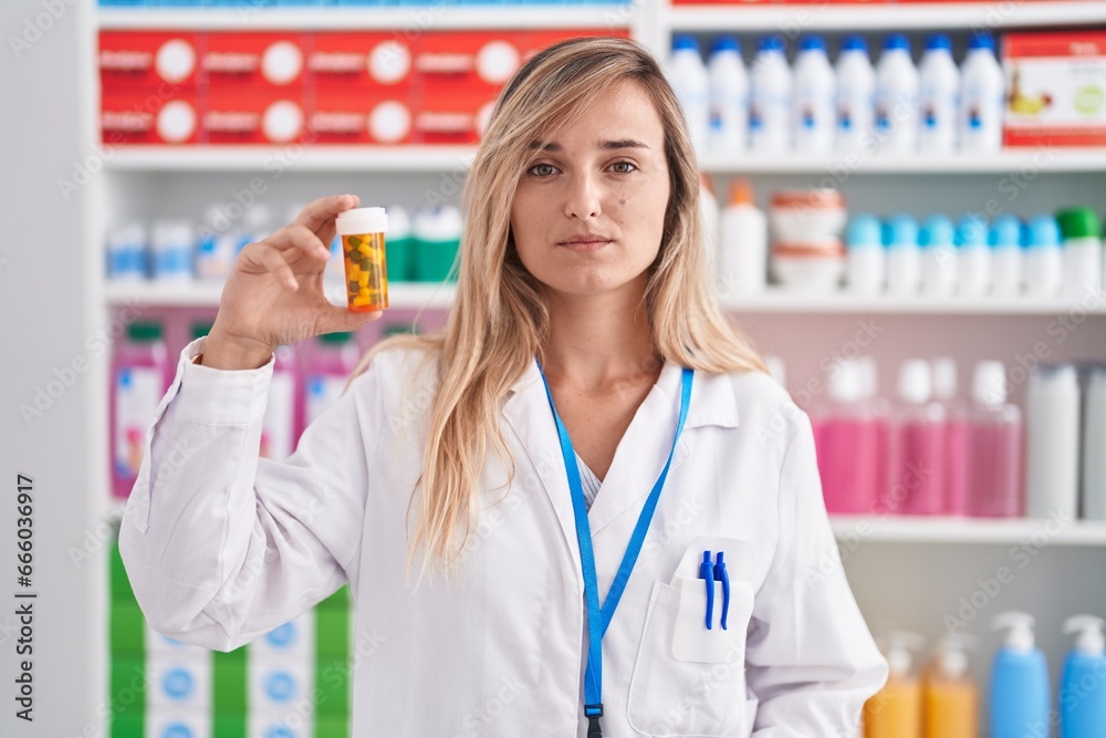 Young blonde woman working at pharmacy drugstore holding pills thinking attitude and sober expression looking self confident