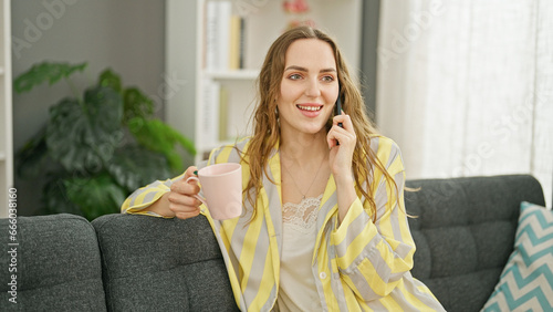 Young blonde woman talking on smartphone drinking coffee at home