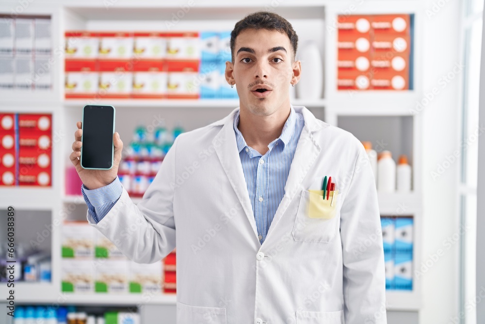 Handsome hispanic man working at pharmacy drugstore showing smartphone screen scared and amazed with open mouth for surprise, disbelief face
