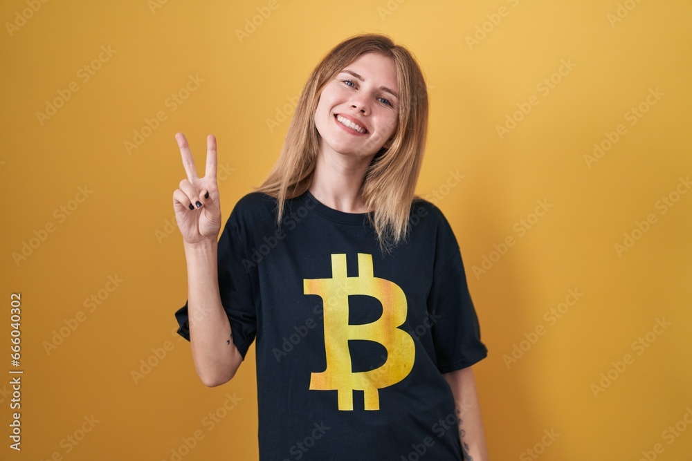 Blonde caucasian woman wearing bitcoin t shirt smiling looking to the camera showing fingers doing victory sign. number two. Blonde caucasian woman wearing bitcoin t shirt smiling looking to the camera showing fingers doing victory sign. number two.
