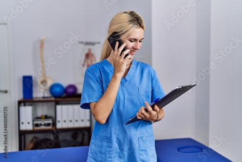 Young blonde woman pysiotherapist talking on smartphone reading document at rehab clinic