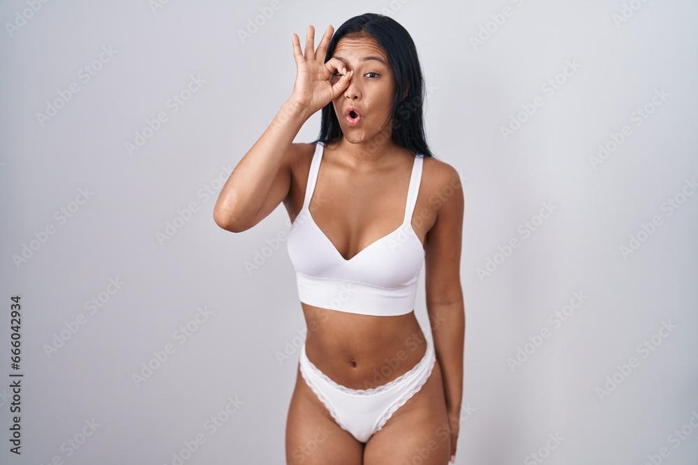 Hispanic woman wearing lingerie doing ok gesture shocked with surprised face, eye looking through fingers. unbelieving expression.