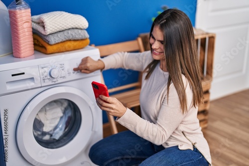Young beautiful hispanic woman turning on washing machine using smartphone at laundry room