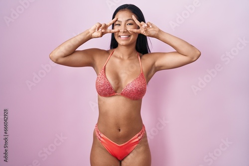 Hispanic woman wearing bikini doing peace symbol with fingers over face, smiling cheerful showing victory © Krakenimages.com