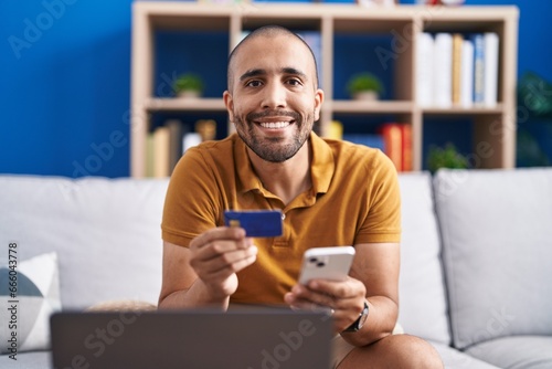 Young latin man using smartphone and credit card sitting on sofa at home