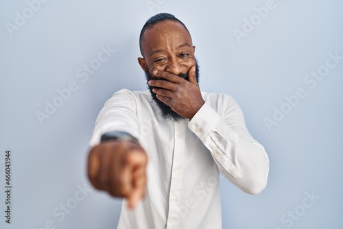 African american man standing over blue background laughing at you, pointing finger to the camera with hand over mouth, shame expression