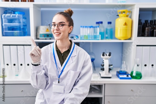 Young hispanic girl working at scientist laboratory smiling with an idea or question pointing finger up with happy face  number one