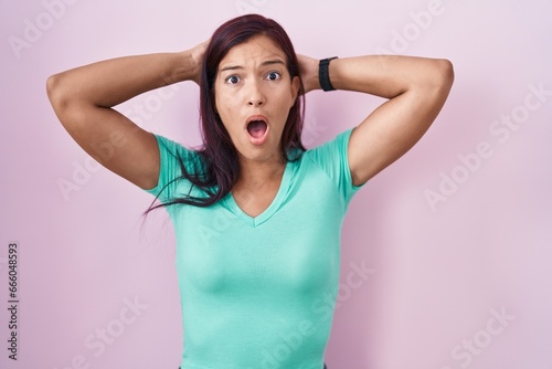 Young hispanic woman standing over pink background crazy and scared with hands on head, afraid and surprised of shock with open mouth