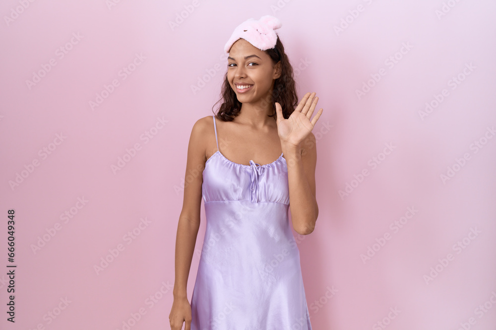 Young hispanic woman wearing sleep mask and nightgown waiving saying hello happy and smiling, friendly welcome gesture Young hispanic woman wearing sleep mask and nightgown waiving saying hello happy and smiling, friendly welcome gesture