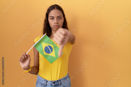 Young hispanic woman holding brazil flag looking unhappy and angry showing rejection and negative with thumbs down gesture. bad expression.