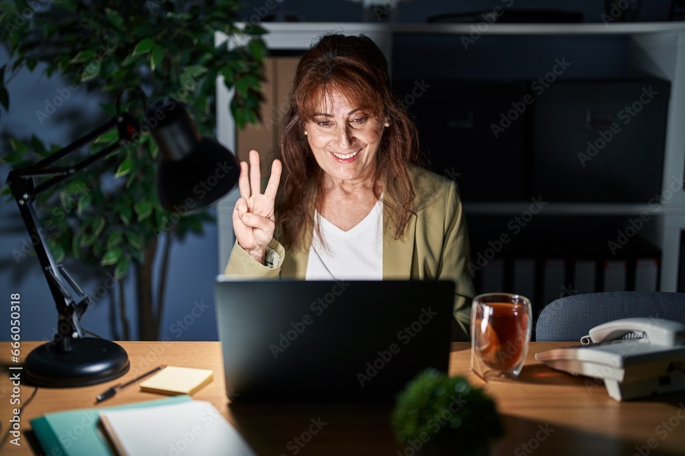 Middle age hispanic woman working using computer laptop at night showing and pointing up with fingers number three while smiling confident and happy. Middle age hispanic woman working using computer laptop at night showing and pointing up with fingers number three while smiling confident and happy.