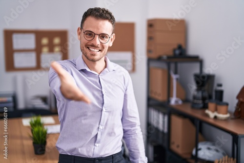 Young hispanic man at the office smiling cheerful offering palm hand giving assistance and acceptance.