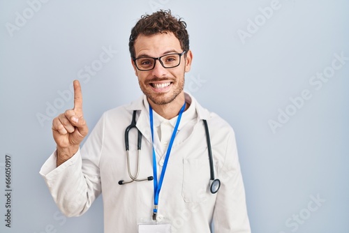 Young hispanic man wearing doctor uniform and stethoscope showing and pointing up with finger number one while smiling confident and happy.