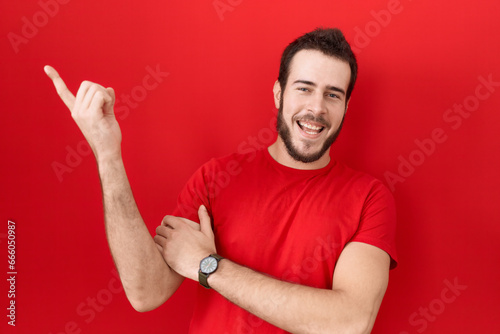 Young hispanic man wearing casual red t shirt with a big smile on face, pointing with hand finger to the side looking at the camera. © Krakenimages.com