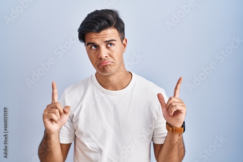 Hispanic man standing over blue background pointing up looking sad and upset, indicating direction with fingers, unhappy and depressed.