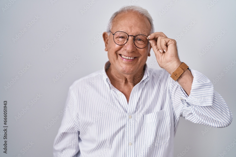 Senior man with grey hair holding glasses with hand looking positive and happy standing and smiling with a confident smile showing teeth