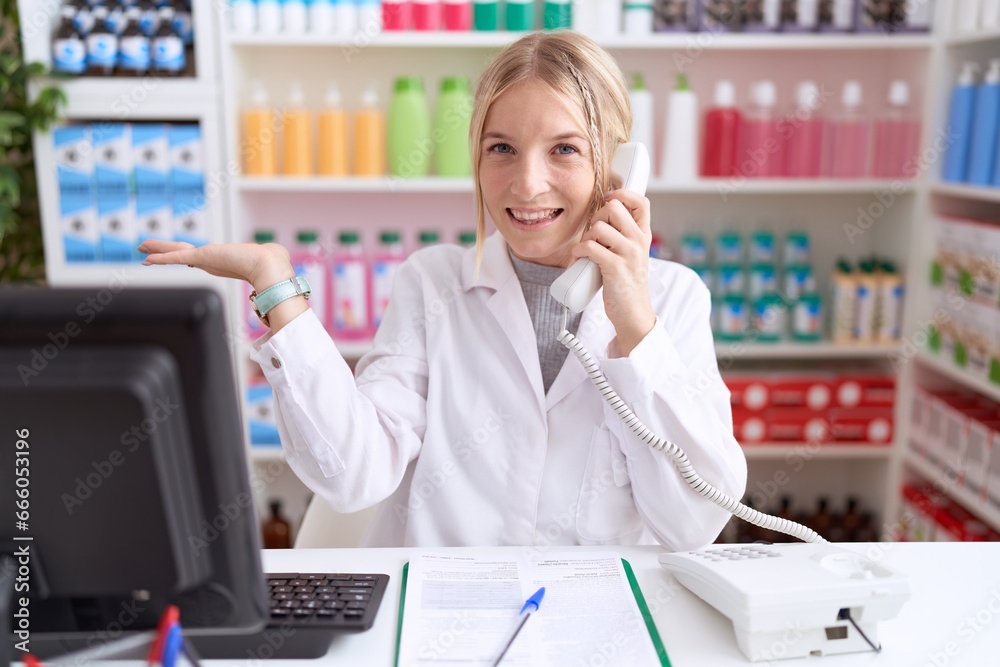 Young caucasian woman working at pharmacy drugstore speaking on the telephone smiling cheerful presenting and pointing with palm of hand looking at the camera.