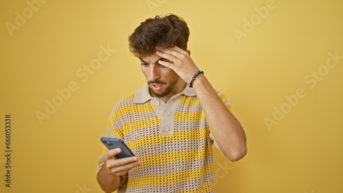 Young arab man, beard and all, amazed and wide-eyed, standing in surprise as he looks at his smartphone, texting away on an isolated yellow wall background. wow, is that an incredible message!