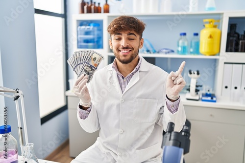 Arab man with beard working at scientist laboratory holding money smiling happy pointing with hand and finger to the side