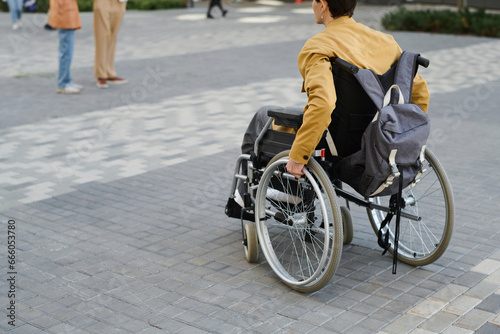 Rear view of young man with disability using wheelchair to move along the street