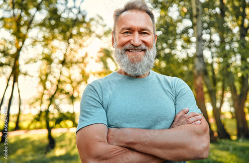 Vitality and active lifestyle. Portrait of fit mature man with hands crossed posing on camera during active training in green forest. Grey-bearded sportsman in blue shirt making confident smile. © HBS