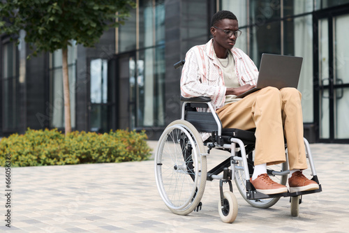 African American man sitting on wheelchair and using laptop outdoors in the city © AnnaStills