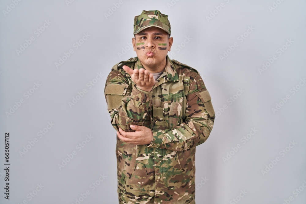 Hispanic young man wearing camouflage army uniform looking at the camera blowing a kiss with hand on air being lovely and sexy. love expression. Hispanic young man wearing camouflage army uniform looking at the camera blowing a kiss with hand on air being lovely and sexy. love expression.