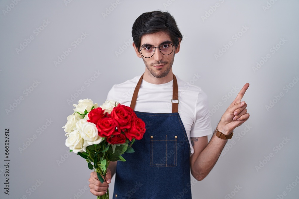 Young hispanic man holding bouquet of white and red roses pointing with hand finger to the side showing advertisement, serious and calm face Young hispanic man holding bouquet of white and red roses pointing with hand finger to the side showing advertisement, serious and calm face
