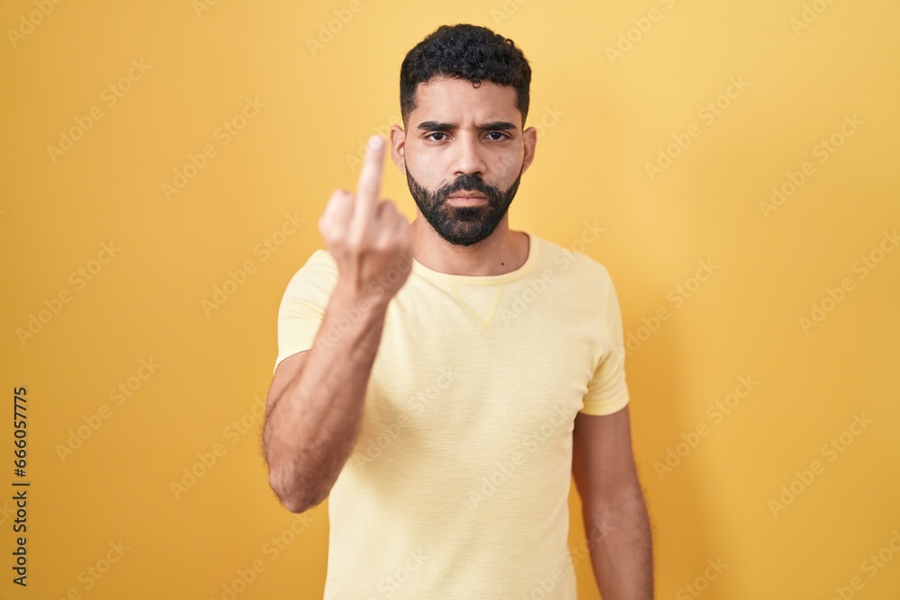 Hispanic man with beard standing over yellow background showing middle finger, impolite and rude fuck off expression Hispanic man with beard standing over yellow background showing middle finger, impolite and rude fuck off expression